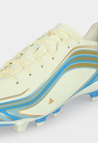 Chaussure de football couleur crème avec des rayures bleues et dorées, laçage à l’avant et semelle à crampons pour jouer en extérieur sur gazon.