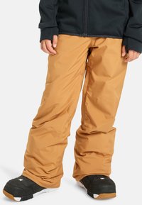 Lyžiarske nohavice v béžovej farbe, vyrobené z odolného materiálu; voľný strih s elastickými manžetami; skombinované s čiernymi snehovými topánkami s bielou podrážkou.