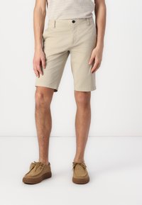 Beige Shorts aus leichtem Material mit flachem Frontdesign, Seitentaschen und einem Knopfverschluss. Kombiniert mit braunen Wildlederschuhen.