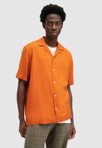 Camisa de manga corta con cuello en brillante tejido naranja, que presenta un cierre de botones en la parte delantera y un sutil bordado en el pecho, combinada con pantalones de cuadros.