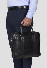 Svart skinnmappe med dobbel håndtak, sølvfarget maskinvare, slankt design og sidelomme med glidelås, holdt av en person kledd i forretningsantrekk.
