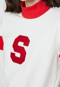 Bílá mikina s vysokým červeným límcem, která má na přední straně velké červené plstěné písmeno "S" a červené pruhy na rukávech.