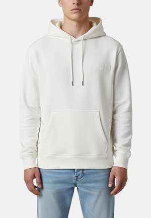 Hombre con sudadera blanca con capucha, bolsillo delantero y logo sutil "4US", combinado con vaqueros azul claro, de pie frente a un fondo liso.