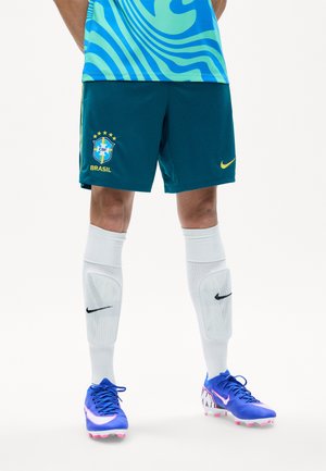 Partie inférieure du corps portant un short de football brésilien, des protège-tibias blancs Nike et des chaussures de football bleues Nike avec des accents roses sur fond blanc.