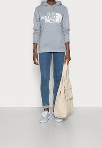 Felpa grigia con logo bianco, jeans skinny blu, sneakers azzurre e borsa beige con tasche frontali. Tessuto liscio, design casual.