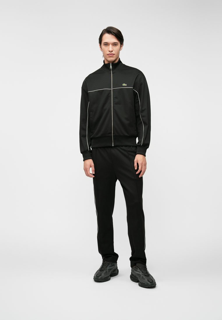 Ensemble de survêtement noir comprenant une veste zippée avec des accents blancs et un pantalon assorti. Tissu lisse avec une coupe ajustée. Porté avec des baskets noires.