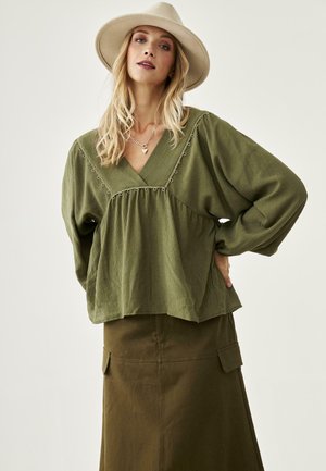 Mujer rubia con sombrero beige, blusa suelta verde oliva con ojales decorativos y falda verde oliva a juego con bolsillos, posando con la mano en la cadera.