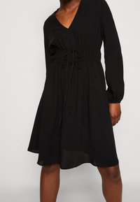 Schwarzes Kleid aus leichtem Stoff, mit V-Ausschnitt, langen Ärmeln, elastischem Taillenbund und einem Kordelzug-Detail.