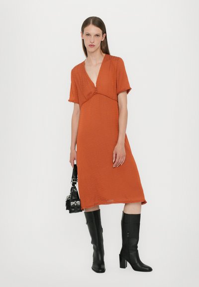 Vestido naranja con escote en V y mangas cortas, que presenta un corte holgado y textura. Combinado con botas de cuero negras hasta la rodilla y un bolso pequeño.