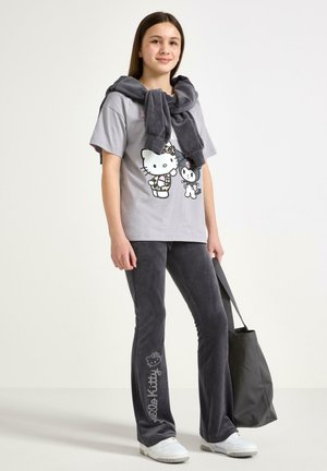 Graues Baumwoll-T-Shirt mit Hello Kitty Grafik, kombiniert mit dunkelgrauen Schlaghosen mit "Hello Kitty"-Text. Das Modell hält eine graue Tragetasche.
