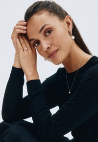 Gioielli in argento, tra cui una delicata collana con una perla, orecchini a perno con perle abbinate e un semplice anello in argento. Abbigliamento con maglione nero.