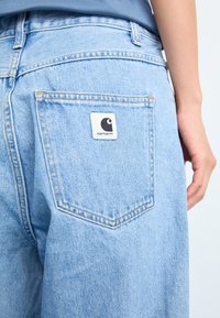 Hellblaue Denim-Jeans verfügen über eine Gesäßtasche mit einem weißen Etikett, auf dem ein schwarzes Logo abgebildet ist. Der Stoff hat eine glatte, leicht abgedreht wirkende Textur.