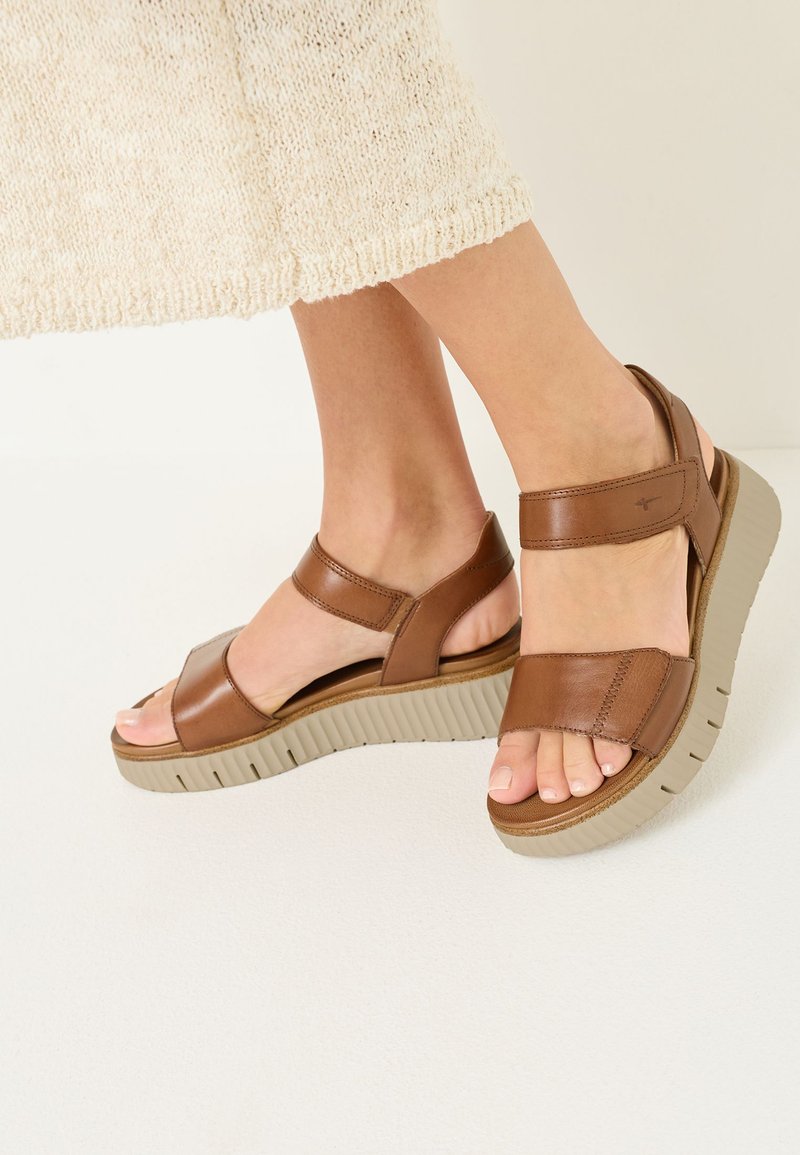 Pies que llevan sandalias de plataforma de cuero marrón con tiras ajustables y suelas gruesas acanaladas beige, mostrados sobre un fondo claro.