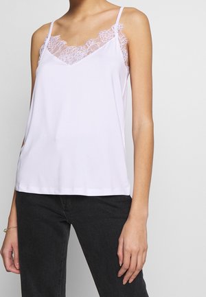 Kobieta ubrana w biały, bezrękawnik z koronkowym wykończeniem, w połączeniu z czarnymi jeansami, stojąca na jednolitym jasnym tle.