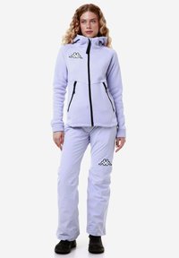 Giacca in pile di colore viola chiaro con colletto alto e zip nere; abbinata a pantaloni viola chiaro con un logo sulla coscia. Stivali neri.