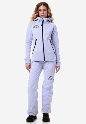 Giacca in pile di colore viola chiaro con colletto alto e zip nere; abbinata a pantaloni viola chiaro con un logo sulla coscia. Stivali neri.