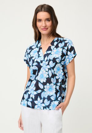 Kobieta o długich brązowych włosach, ubrana w czarną bluzkę z krótkim rękawem z dużym niebiesko-białym kwiatowym wzorem oraz białe spodnie, lekko uśmiechnięta.