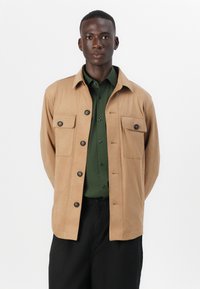 Veste button-up beige avec deux poches poitrine, faite d'un tissu lisse. Associée à une chemise verte, un pantalon noir, coupe slim, au design structuré.
