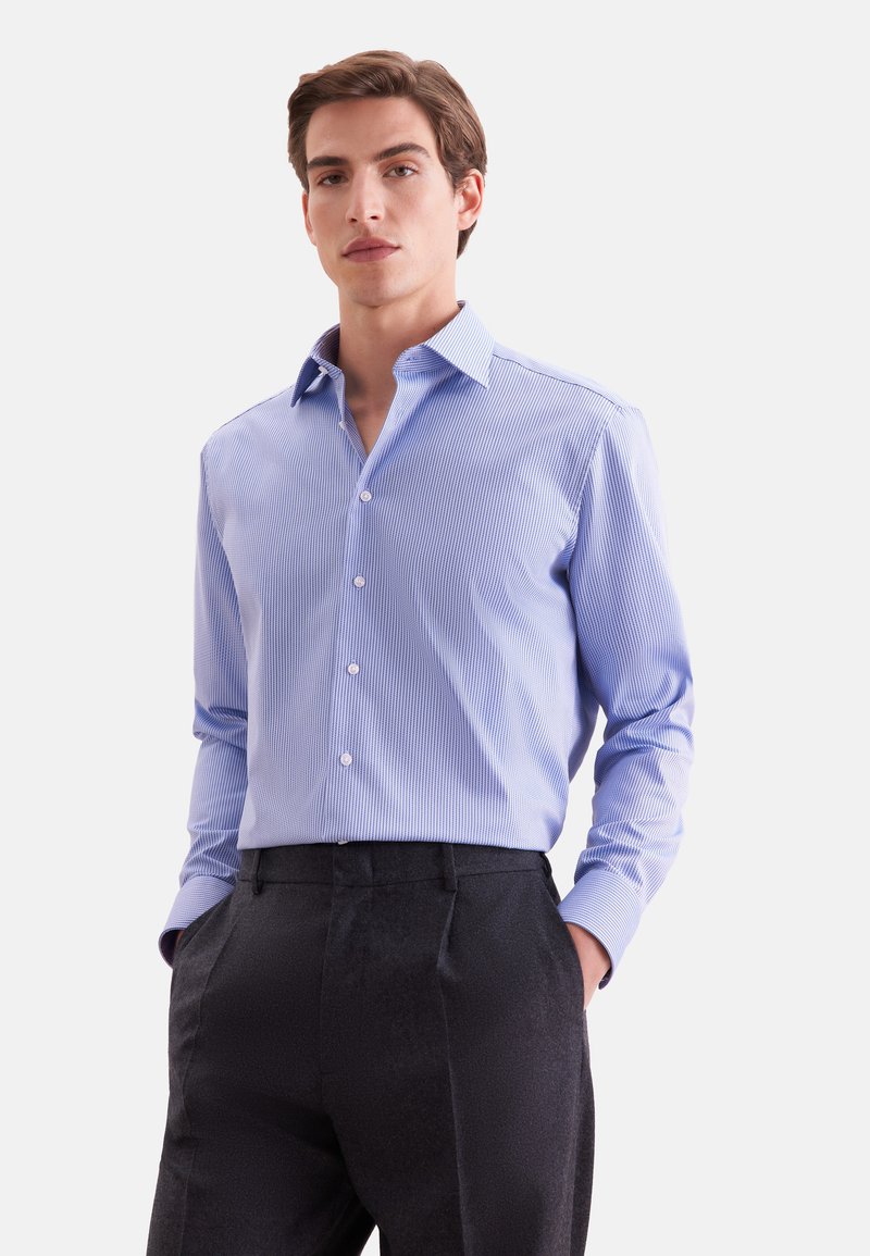 Jeune homme portant une chemise habillée bleu clair à rayures avec un pantalon noir, debout les mains dans les poches devant un fond blanc uni.
