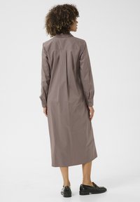 Ilga ruda marškinėlių suknelė, pagaminta iš lengvos medžiagos, turinti apykaklės iškirptę, mygtukų rankoves ir tiesią siluetą. Juodi batai.