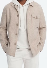 Beige Jacke aus Wollmischung mit Kragen, Vorderknöpfen und einer Brusttasche. Trägt man über einem hellgrauen Hoodie.