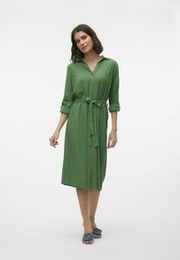 Abito verde, con bottoni, realizzato in tessuto morbido, con cintura legata in vita, maniche lunghe arrotolate fino ai gomiti e vestibilità rilassata.