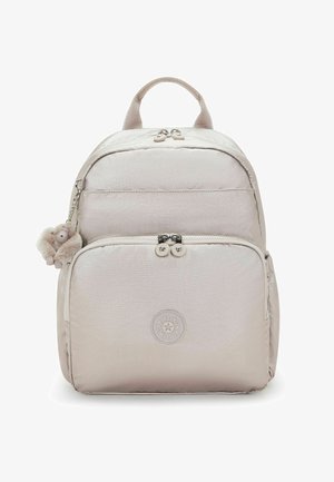 Mochila beige claro con bolsillo delantero con cremallera, doble tirador de cremallera, parche de logo redondo y un pequeño llavero de mono de peluche colgando del costado.