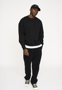 Sort sweatshirt med ribbede manchetter og kant, lagd over en hvid t-shirt; parret med sorte bukser og sorte sneakers med hvide såler.