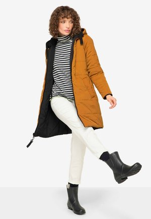 Quilted amber parka with black lining, striped navy and white turtleneck, cream pants, and black ankle boots on a white background.