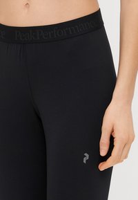 Leggings atléticos pretos com uma cintura alta, apresentando um discreto logotipo da Peak Performance em cinza. Tecido suave e ajuste justo.
