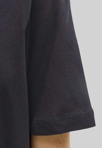 Tela negra de manga corta en el brazo contra un fondo liso gris claro, mostrando el detalle de costura en el dobladillo.