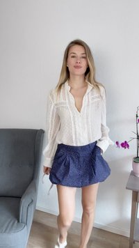 Witte blouse met gestructureerde accenten en een V-hals, gecombineerd met een marineblauwe skort met witte stippen. Kamer met een plant en een grijze stoel.