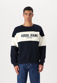 Navy en crèmekleurige color-blocked sweatshirt met de tekst "GUESS JEANS", een relaxte pasvorm, ronde halslijn en geribde mouwen. Draag het met blauwe jeans.