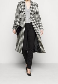 Abrigo largo de cuadros blanco y negro con grandes solapas, combinado con una blusa gris de botones, pantalones negros y tacones altos negros.