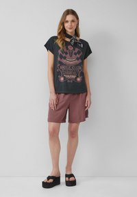Zwart graphic t-shirt met roze print, korte mouwen, gecombineerd met lichtbruine shorts. Zwarte platform sandalen hebben een gedraaide banddetail.