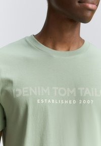 Svetlozelené bavlnené tričko s klasickým výstrihom a potlačou textu "DENIM TOM TAILOR ZALOŽENÉ 2007" v kontrastnej farbe.