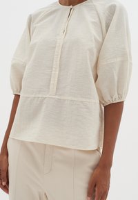 Blusa color crema dalla vestibilità morbida, con scollatura rotonda e maniche arricciate. Dettaglio frontale plissettato e chiusura con bottone al collo.