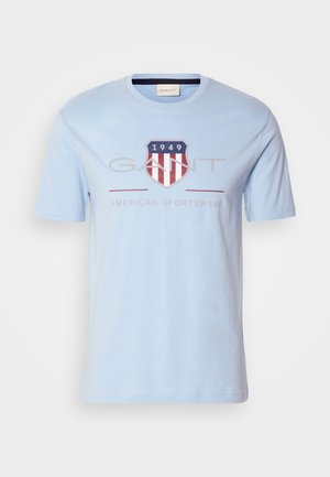 Helehelesina sinine puuvillane T-särk, millel on kilbi logo punaste ja valgete triipudega, "1949" ning tekst "GANT AMERICAN SPORTSWEAR." Lühikesed varrukad.