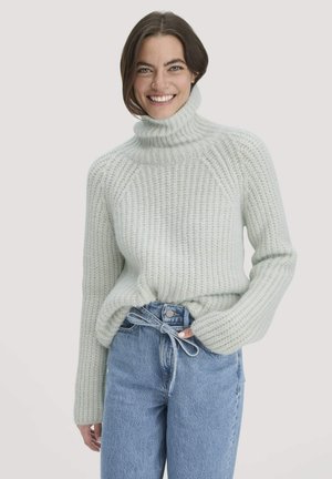 Woman smiling, wearing a light gray chunky knit turtleneck sweater and high-waisted blue jeans with a tied belt loop detail.