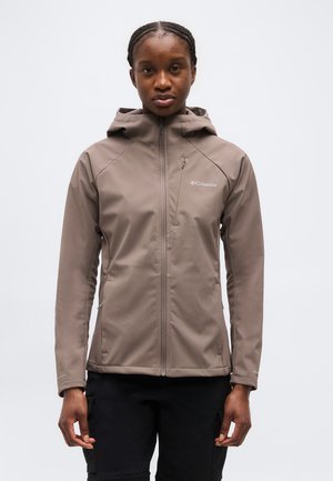 Woman wearing a brown Columbia hooded jacket zipped up, standing facing forward against a plain background.