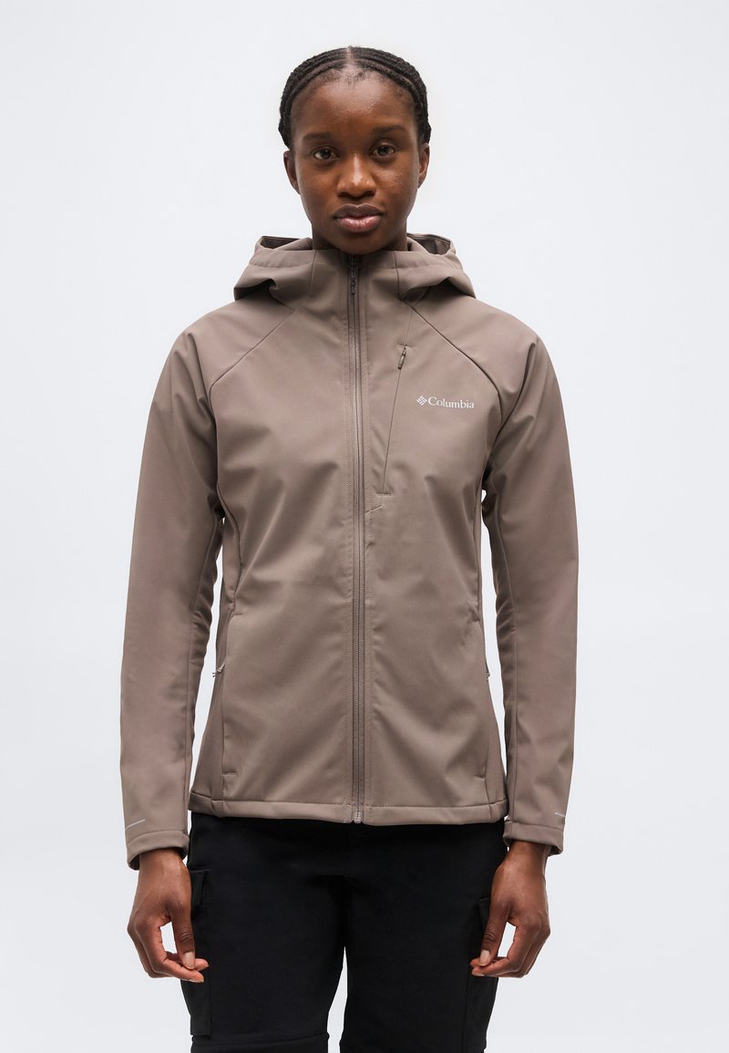 Femme portant une veste à capuche Columbia marron, zippée, debout face à l'avant sur un fond uni.