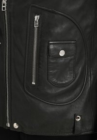 Veste en cuir noir avec une poche zippée, une poche à rabat arrondie avec un bouton-pression métallique, et des détails de couture visibles. Texture lisse.