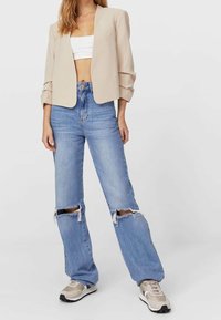 Blazer cropped beige con maniche arrotolate, abbinato a jeans chiari strappati con strappi alle ginocchia, e sneakers atletiche beige.