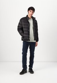 Svart pufferjakke med høy krage, trykknapper og en teksturert overflate, båret over en lys grønn genser og mørke jeans.