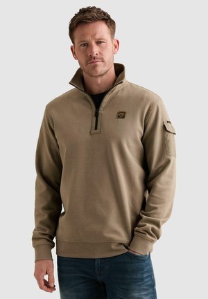 Homme portant un sweat-shirt beige à fermeture éclair quart, avec un petit patch sur la poitrine et une poche zippée sur la partie supérieure de la manche gauche, associé à un jean bleu.