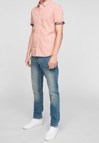 Růžová košile s krátkým rukávem a vzorovanými manžetami, kombinovaná s světlými modrými džínami a bílými teniskami. Ležérní střih a viditelná textura látky.
