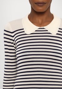 Osoba v krémovém a námořnickém pruhovaném svetru se vlnitým bílým límcem Peter Pan, zachycena od ramen k ústům.