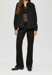 Schwarze Cordjacke mit Knopfleiste und zwei Brusttaschen, getragen über einem schwarzen Spitzentop und kombiniert mit schwarzen Schlaghosen.
