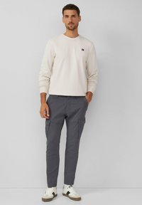 Sudadera de un tono beige claro con puños acanalados y un pequeño logo, combinada con pantalones cargo grises que tienen bolsillos laterales, y zapatillas blancas con detalles en negro.