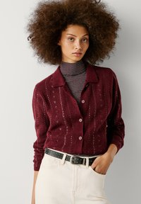 Μπορντώ πλεκτό κασκόλ με ραβδωτή υφή, κλείσιμο με κουμπιά και διακριτές λαμπερές λεπτομέρειες. Συνδυάζεται με ένα ριγέ τζέρσεϊ και μαύρη ζώνη.